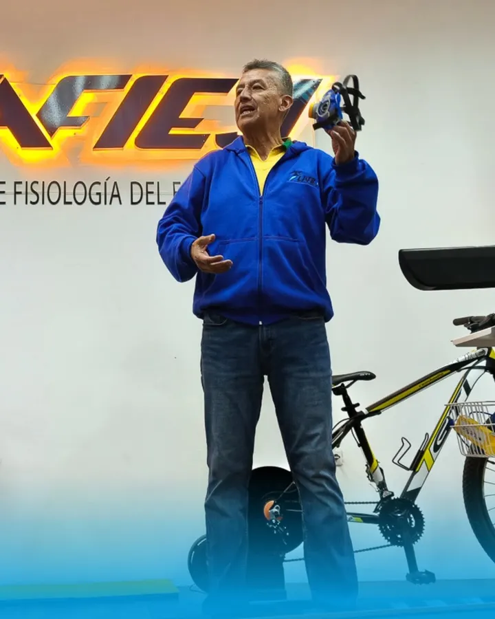 El fundador de LAFIEJ sostiene en alto la máscara de ventilometría junto a una bicicleta de montaña, frente al rótulo retroiluminado