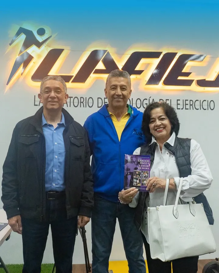 Tres personas sonríen bajo el rótulo retroiluminado de LAFIEJ; el hombre al centro sostiene el libro «Ejercicio Físico y Cáncer de Mama»
