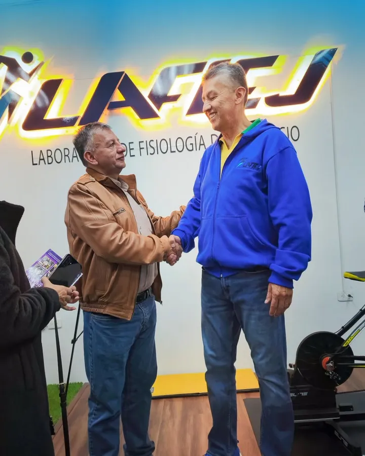 Dos hombres se dan la mano durante la inauguración de LAFIEJ, bajo el rótulo tridimensional retroiluminado con halo ámbar; a la derecha, una bicicleta de ruta montada sobre un rodillo de entrenamiento